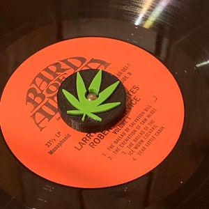 May include: A red vinyl record with the text "Bard of Avon" and "Larry Roberts Volume I" on the label. The record is 33 1/3 RPM and monophonic. The record is on a turntable with a green marijuana leaf-shaped record weight.