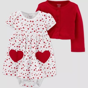 May include: A white baby dress with a red heart pattern and two large red felt hearts on the front. The dress is paired with a red cardigan with a button closure. The text "Just One You" is printed on the dress and cardigan.