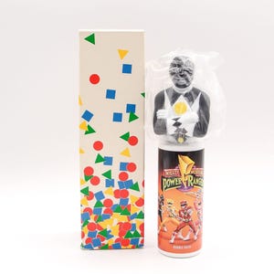 May include: A Mighty Morphin Power Rangers bubble bath container featuring a black ranger figure topper encased in plastic. The cylindrical bottle displays colorful character graphics and the show logo. Beside it stands a tall, rectangular box decorated with a pattern of small red, blue, and yellow geometric shapes.