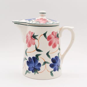 May include: A cream-colored ceramic pitcher with a lid, featuring a hand-painted floral pattern of pink and blue flowers with green leaves and thin orange vines. The pitcher has a curved handle, a wide spout, and a matching lid with a small knob.