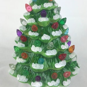 May include: A ceramic Christmas tree night light with a green base and white snow. The tree is decorated with colorful light bulbs. The night light has a white plug and an on/off switch.