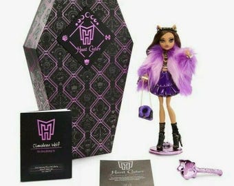 May include: A Monster High Haunt Couture doll with a purple faux fur coat, skirt, and purse. The doll has brown hair and is standing on a round base. The set includes a coffin-shaped box, a booklet, and a key.