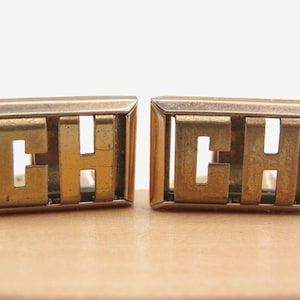 May include: A pair of vintage gold-toned cufflinks. Each cufflink features the letters "CH" in a rectangular frame. The letters are cut out, creating negative space. The cufflinks are displayed on a light brown surface.
