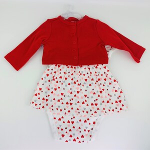 May include: A red cardigan with a white button closure over a white onesie with a pink and red heart print. The onesie has a skirt overlay with the same heart print.