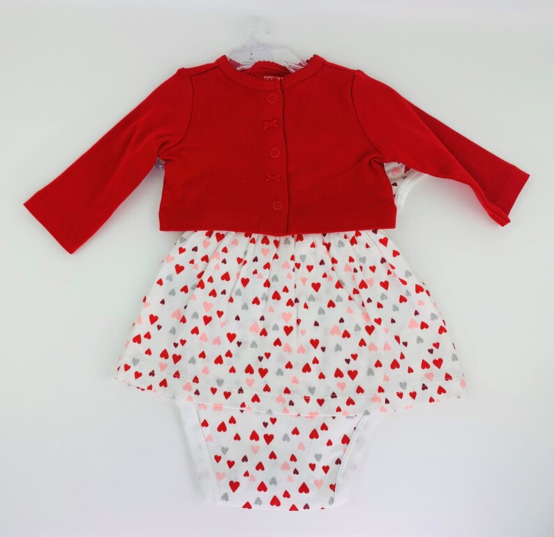 May include: A red cardigan with a white button closure over a white onesie with a pink and red heart print. The onesie has a skirt overlay with the same heart print.