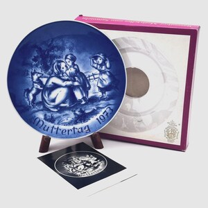 May include: A blue and white decorative plate with a mother and children scene, the words "Muttertag 1973" are printed on the plate. The plate is displayed on a wooden stand, with a matching box in the background.