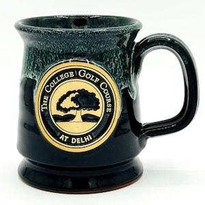 May include: A ceramic mug with a dark green and black glaze. The mug features a gold emblem with the text "THE COLLEGE GOLF COURSE AT DELHI" and a tree graphic. The handle is a rounded, dark green color.