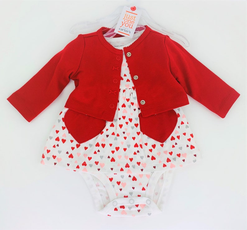 May include: A red cardigan with a white bodysuit and a white dress with a heart pattern. The dress has red heart accents. The tag says "Just One You Carter's".
