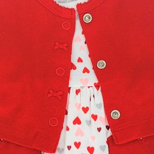 May include: Red cardigan with a white dress underneath. The dress has a heart pattern with red, pink, and silver hearts.