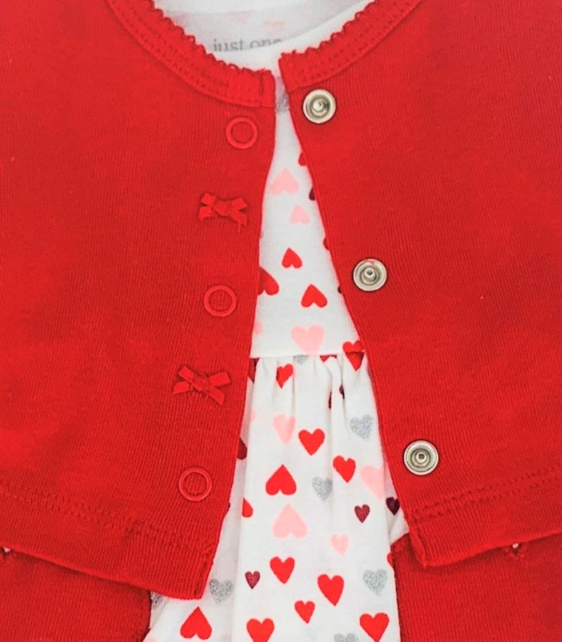 May include: Red cardigan with a white dress underneath. The dress has a heart pattern with red, pink, and silver hearts.