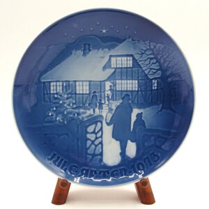 May include: A blue and white decorative plate featuring a winter scene with a house, people, and a Christmas tree. The plate has the text "JUL. AFTEN 1973" and is displayed on a wooden stand. A star is in the sky.