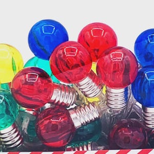 May include: A collection of colorful, round Christmas light bulbs. The bulbs are in various colors, including red, blue, green, and yellow. The bulbs have silver bases and are arranged in a pile.