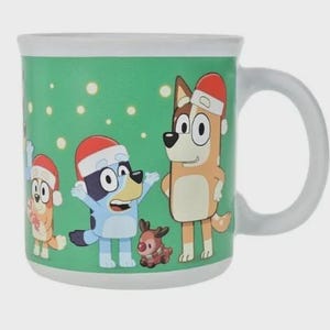 May include: White ceramic mug featuring characters from the animated series Bluey, wearing Santa hats. The mug has a green background with white dots and a white handle. The characters include Bluey, Bingo, and a reindeer.