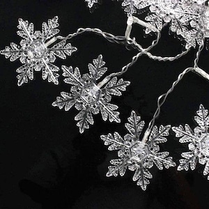 May include: A string of clear, snowflake-shaped LED lights. Each snowflake has a faceted center and detailed, leaf-like edges. The lights are connected by a thin, clear wire, creating a festive, wintery aesthetic.