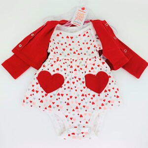 May include: A white baby onesie with a red heart print and two large red felt hearts. The onesie is part of a three-piece outfit that includes a red cardigan and a white dress with a red heart print. The outfit is labeled "Just One You Carter's".