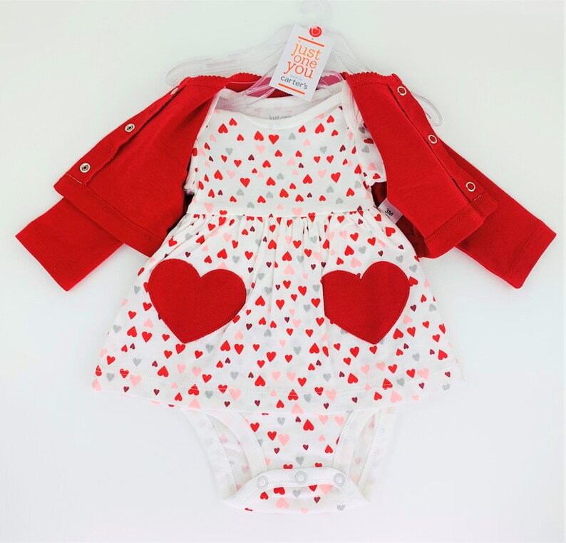 May include: A white baby onesie with a red heart print and two large red felt hearts. The onesie is part of a three-piece outfit that includes a red cardigan and a white dress with a red heart print. The outfit is labeled "Just One You Carter's".