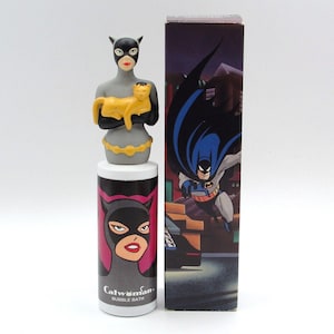 May include: A Catwoman bubble bath container featuring a gray bust of the character holding a yellow cat, atop a cylindrical bottle with a pink and black graphic of Catwoman's face and the text Catwoman BUBBLE BATH, next to a tall box.