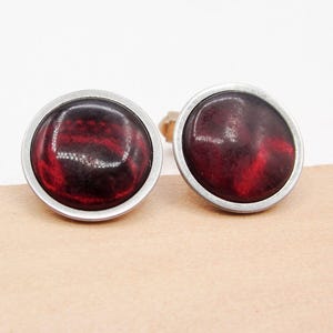 May include: A pair of round cufflinks with a dark red, marbled design. Each cufflink has a silver-colored metal border. The cufflinks are displayed on a light brown surface.