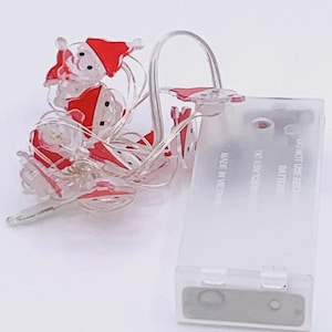 May include: String of Christmas lights featuring small, clear Santa Claus figures with red hats and white beards. The lights are connected to a clear battery pack. The battery pack has the text "MADE IN VIETNAM".