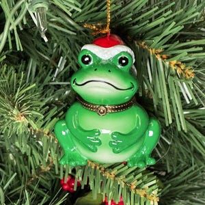 May include: A green frog Christmas ornament with a Santa hat and a gold-colored collar. The frog is smiling and has black eyes. The ornament is hanging on a Christmas tree.