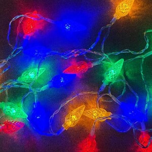 May include: A string of colorful Christmas lights with red, blue, green, and yellow bulbs. The lights are on a clear wire and are arranged in a tangled pattern. The bulbs have a textured, faceted design.