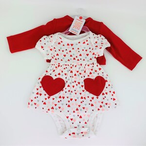 May include: A white baby onesie with a red heart print and two large red hearts on the front. The onesie is layered over a red long-sleeve shirt with the text "Just One You Carter's" on the tag.