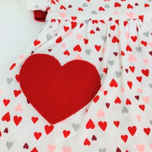 May include: White dress with a red heart applique and a pattern of red, pink, and silver hearts.