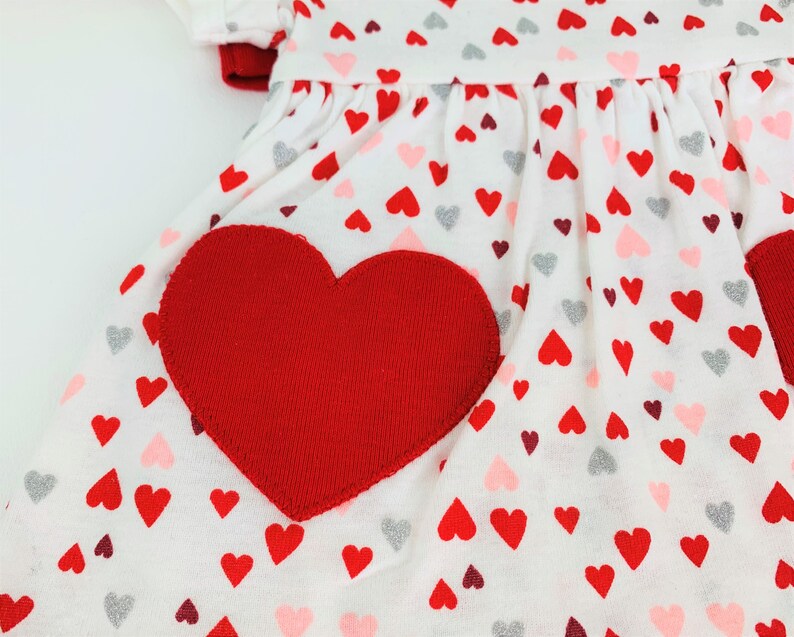 May include: White dress with a red heart applique and a pattern of red, pink, and silver hearts.