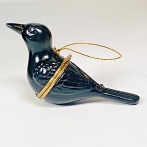 May include: A glossy black bird-shaped trinket box with a gold-colored band around its middle. The bird has a long beak and a gold string for hanging. The trinket box is a decorative item.