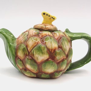May include: A ceramic teapot shaped like an artichoke. The teapot is primarily green with brown and tan accents, and a yellow stem on the lid. The handle and spout are also green. A unique and decorative kitchen item.