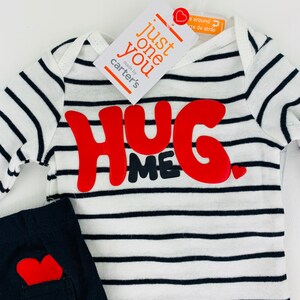 May include: A white and black striped long-sleeve shirt with red felt letters that say "Hug Me" and a small red heart. The shirt is paired with black pants with a red heart on the pocket. The shirt tag says "Just one you" and "made by Carter's".