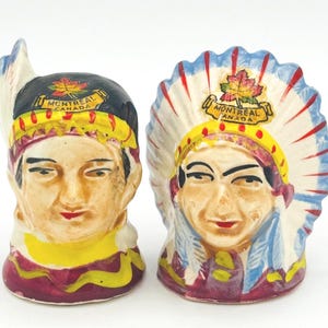 Può includere: Due statuine in ceramica raffiguranti figure di nativi americani, una con un copricapo e l'altra con un copricapo di piume. Entrambe presentano la scritta "MONTREAL CANADA" e un emblema a forma di foglia d'acero. Le statuine sono dipinte con colori vivaci.