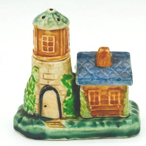 May include: A ceramic lighthouse and cottage salt and pepper shaker set. The lighthouse is tan with a brown top and windows. The cottage has a blue roof and brown walls. The base is green with a grassy design.