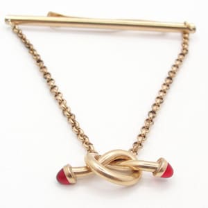 May include: A gold-toned tie bar featuring a horizontal bar connected by two delicate link chains to a central knot-shaped charm. The charm is accented with small, smooth, red, conical-shaped tips on each end of the knot structure.