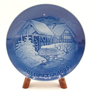 May include: Blue and white ceramic plate with a winter scene. The plate features a snow-covered house, trees, and the text "Jule Aften 1975". The plate is displayed on a wooden stand.