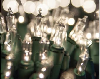 May include: Close-up of a string of clear mini Christmas lights with green wire. The lights are illuminated, creating a warm, soft glow. The background is blurred, with out-of-focus light orbs.