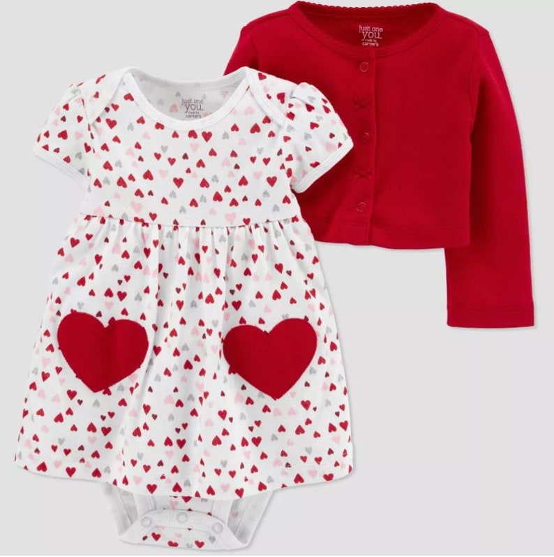 May include: A white baby dress with a red heart pattern and two large red felt hearts on the front. The dress is paired with a red cardigan with a button closure. The text "Just One You" is printed on the dress and cardigan.
