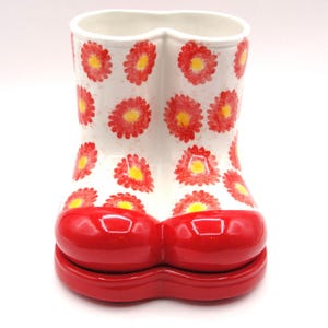 May include: A ceramic vase shaped like a boot, featuring a white base adorned with red and yellow daisy patterns. The boot's toe and base are a glossy red. The vase has a unique, playful design.