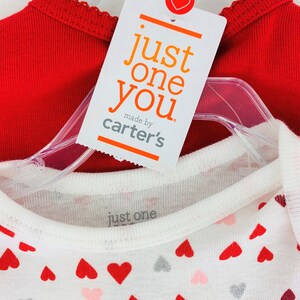 May include: Two red and white shirts with a heart pattern. The red shirt has a scalloped neckline. The white shirt has a small tag that says "just one" and a larger tag that says "just one you made by carter's".