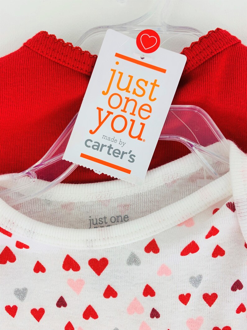 May include: Two red and white shirts with a heart pattern. The red shirt has a scalloped neckline. The white shirt has a small tag that says "just one" and a larger tag that says "just one you made by carter's".