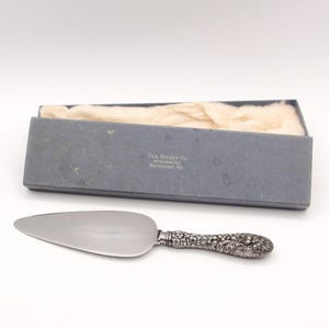 May include: A silver cake server with a smooth, pointed blade and a handle featuring an intricate floral relief pattern. The server is accompanied by a rectangular blue storage box with the text The Stieff Co. Silversmiths Baltimore MD.