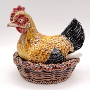 May include: A ceramic hen figurine with a red comb and wattle, perched atop a brown basket. The hen has yellow, brown, and black feathers. A white egg is visible in the basket. Decorative kitchenware.