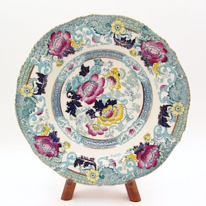 Puede incluir: Un plato de cerámica decorativo con borde festoneado, con un diseño floral en rosa, amarillo y azul. El plato se exhibe sobre un pequeño soporte de madera, mostrando sus intrincados patrones y colores. El diseño del plato incluye un arreglo floral central y un borde detallado.