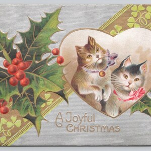 May include: Vintage Christmas postcard featuring two kittens inside a heart-shaped frame. The postcard has holly and berries, a gold ribbon with shamrocks, and the text "A Joyful Christmas". The background is silver.