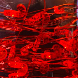 May include: A close-up of a clear container filled with red, glowing LED lights and copper wire. The lights are shaped like candy canes and are scattered throughout the container. The wire is intertwined, creating a festive and decorative display.