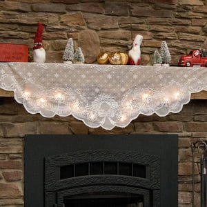 May include: White lace fireplace mantel scarf with a scalloped edge and a garland design. The scarf is decorated with small white lights and is draped over a brick fireplace.