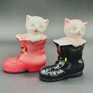 May include: Two ceramic cat figurines, each emerging from a boot. One cat is white with green eyes and sits in a red boot. The other cat is pink with green eyes and sits in a black boot. The boots have a glossy finish.