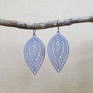 May include: A pair of light periwinkle blue leaf-shaped earrings. The earrings have a delicate, openwork design with intricate patterns and small circular cutouts. They hang from black metal hooks and are displayed against a neutral background.