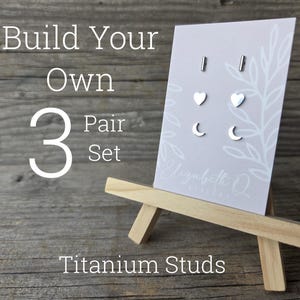 May include: A display of titanium stud earrings on a small wooden easel. The earrings are in the shapes of bars, hearts, and crescent moons. The text on the display reads "Build Your Own 3 Pair Set Titanium Studs."