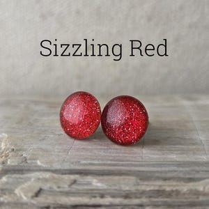 May include: A pair of round, red stud earrings with a glittery finish. The earrings are displayed on a textured, gray surface. The text "Sizzling Red" is above the earrings.